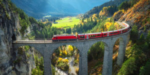 Aerial view of modern red train on Landwasser viaduct in alpine mountains, colorful forest at sunset in autumn. Bernina Express, Switzerland. Top view of train in Alps, railroad, green trees in fall